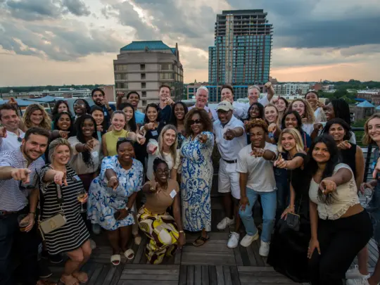 A group of students, professors and alumni smiling and pointing at the camera at a rooftop restaurant during the sunset in Durham, NC